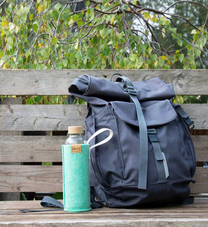 Minzfarbene Filzhülle für Glastrinkflasche mit Trageschlaufe steht auf Bank neben einem Rucksack